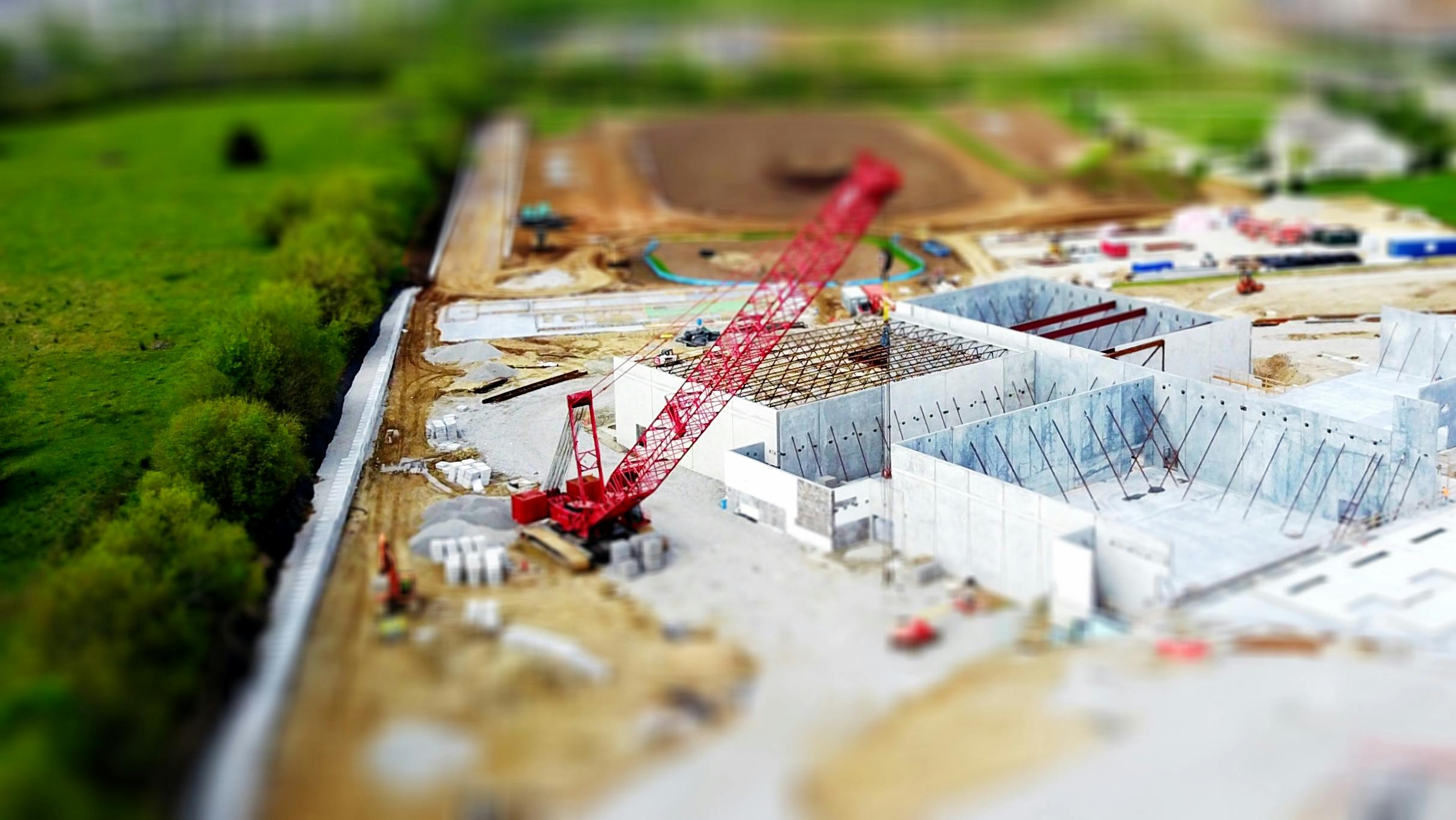 Aerial view of a construction site with a red crane amidst green fields and concrete buildings.